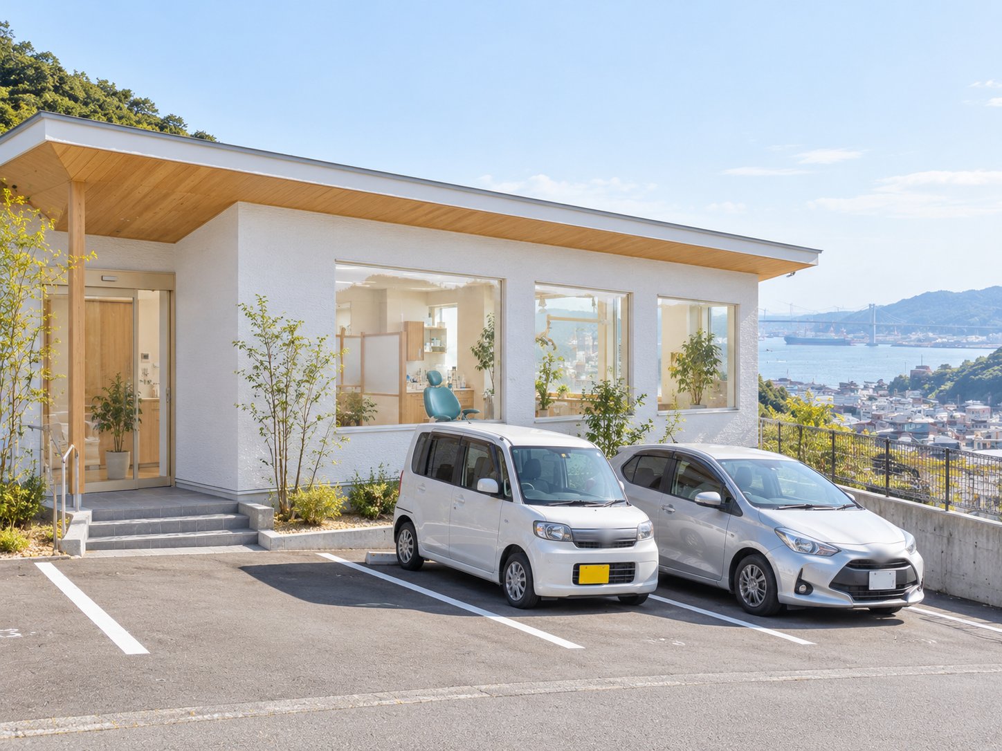 坂の上に建つ白い平屋の医院の外観と、建物の前の駐車場。奥に海と町並みが見える